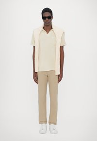 Lichtbeige polo-shirt met zak, crèmekleurige kabelgebreide vest, tan broek en witte sneakers. Model draagt donkere zonnebril. Eenvoudig, strak ontwerp.