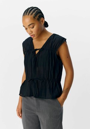 Top negro con pliegues, mangas cortas, escote en V y detalle de cintura anudada. Combinado con pantalones grises, el tejido parece ligero y con textura.