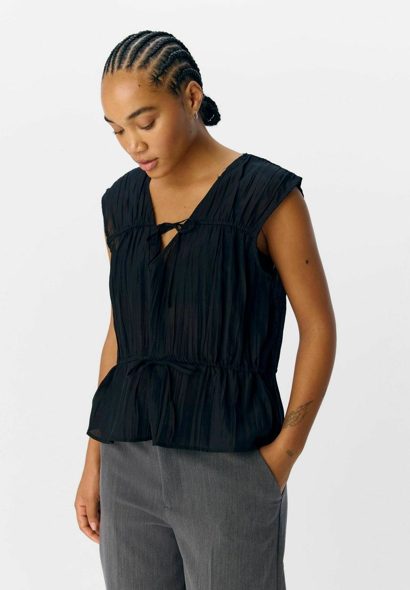 Top negro con pliegues, mangas cortas, escote en V y detalle de cintura anudada. Combinado con pantalones grises, el tejido parece ligero y con textura.