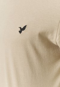 Beige cotton T-shirt featuring a black embroidered bird logo on the left chest area. Smooth texture, regular fit with short sleeves.