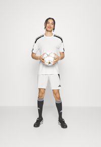Vit idrottströja och shorts med svarta accenter, svarta strumpor med vita ränder, och svarta fotbollsskor; håller i en mönstrad fotboll.