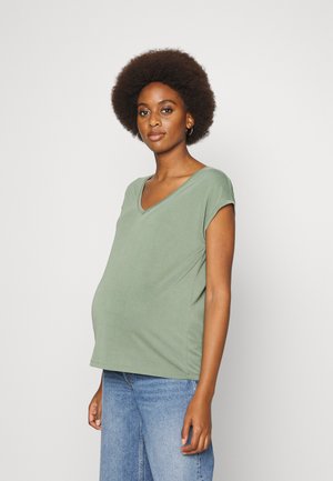 Camiseta de maternidade em verde suave, com mangas curtas, decote em V, textura suave, combinada com jeans de ganga, apresenta um corte solto na área do abdómen.