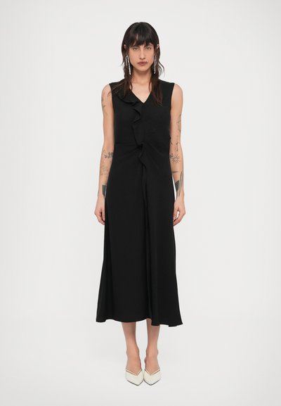 Woman stands wearing a sleeveless black midi dress with a ruffle detail down the front and white pointed-toe heels on a plain background.
