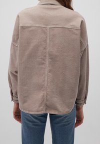 Camicia in velluto a coste di colore marrone chiaro, caratterizzata da una vestibilità rilassata, polsini con bottoni e una texture a coste verticali. La vista posteriore mostra dettagli delle cuciture.