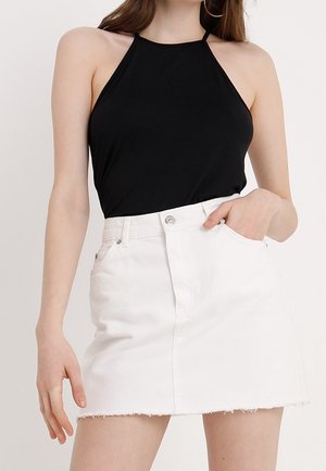 Vrouw in een mouwloze zwarte haltertop, gestoken in een witte denim minirok met rafelige rand, poseert met één hand in de zak.