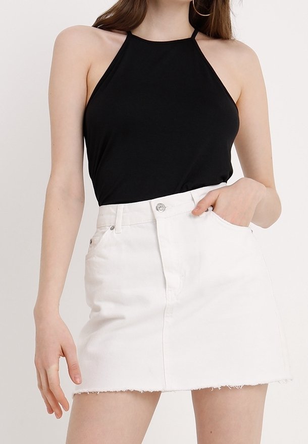 Kvinna i en ärmlös svart halterneck-top som är inskjuten i en vit denim mini kjol med fransar, poserar med en hand i fickan.