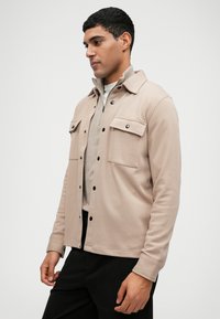 Giacca marrone chiaro realizzata in tessuto morbido, con chiusura a bottoni, due tasche sul petto e design con colletto. Indossata sopra un top grigio.