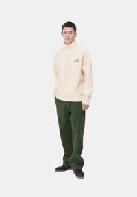 Sudadera con cremallera de color beige claro, con logo bordado, combinada con pantalones verde oscuro y zapatos negros, con un corte holgado y textura suave.