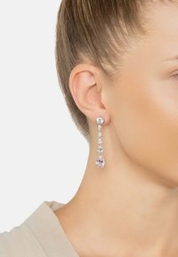 Primo piano di un orecchio femminile che indossa un lungo orecchino pendente con gemme a goccia trasparenti e rosa.