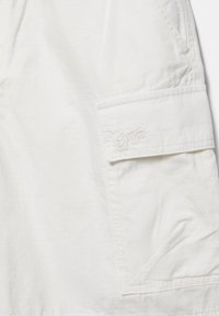 Biały materiał z przednią klapą na kieszeń typu cargo, ozdobioną przeszyciami i subtelnym haftowanym logo.