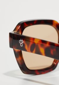 Tortoiseshell sunglasses with square frames, brown lenses, and a glossy finish. Features a logo on the temple and chunky design.