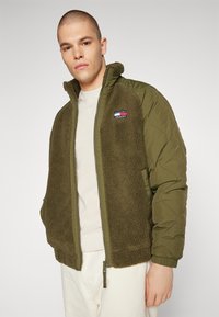Chaqueta verde olivo con un frente de forro polar texturizado, mangas acolchadas y cierre de cremallera. Pequeño parche de logo en la parte superior izquierda.