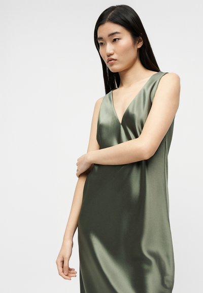 Νεαρή γυναίκα με μακριά σκούρα μαλλιά που φοράει αμάνικο σατέν φόρεμα ελιάς με V-λαιμόκοψη, σταυρωμένα χέρια, κοιτάζει προς τα κάτω με σκέψη.