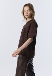 T-shirt oversize en suède marron avec des manches courtes, un col rond et une broderie texturée sur le devant, associé à un pantalon sombre à motifs.