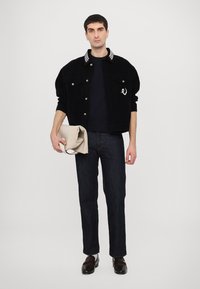 Giacca di velluto a coste nera con colletto a righe, T-shirt scura e jeans di denim scuro. Tiene in mano una borsa a tracolla beige. Scarpe marroni.
