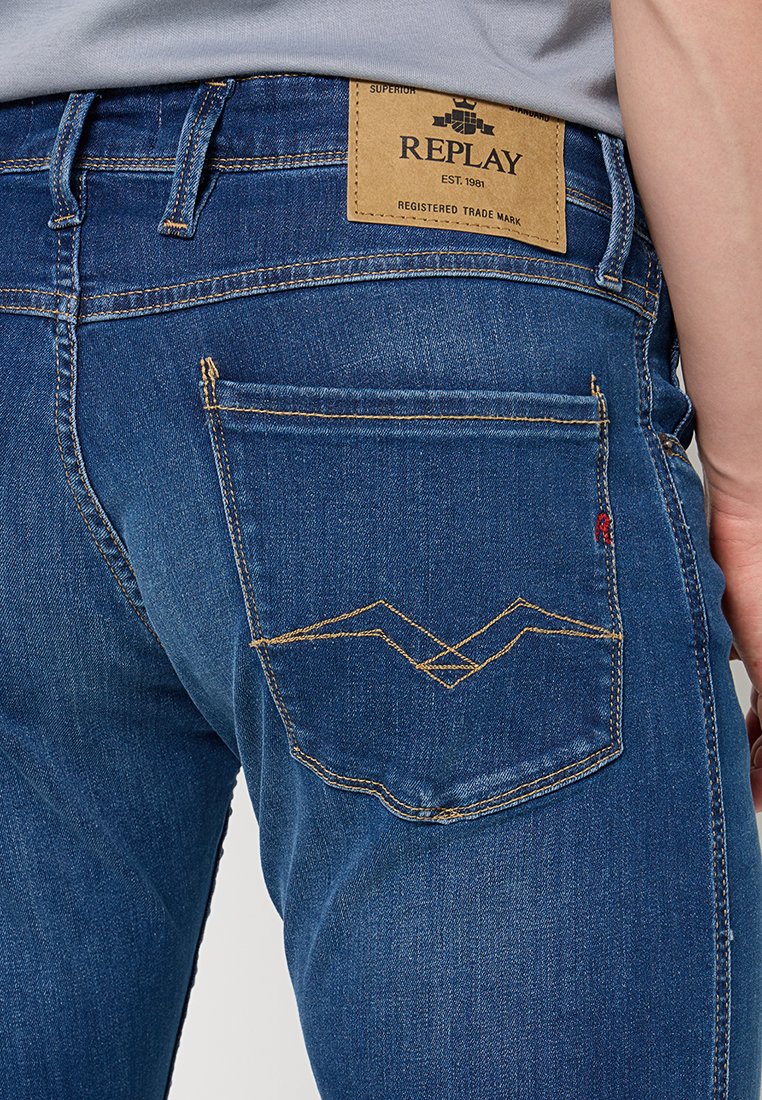 Jeans dżinsowe w ciemnym niebieskim kolorze o prostym kroju. Posiadają żółte przeszycia na tylnej kieszeni oraz prostokątną, tkaną metkę z logo 'REPLAY'.