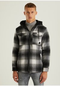 Jeune homme portant une veste zippée à carreaux noirs et blancs avec une capuche par-dessus un pull à col roulé gris, debout devant un fond uni.