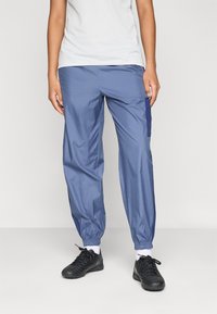 Personne portant un pantalon de jogging bleu avec des poignets élastiques, des chaussures de sport noires et un haut blanc à manches courtes, se tenant sur un fond uni.
