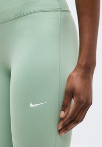 Main à peau foncée reposant à côté d'un legging Nike vert clair avec un logo swoosh blanc sur la cuisse, sur un fond clair uni.