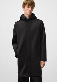 Manteau noir à capuche avec une texture lisse, présentant des poches latérales et un design minimaliste. Le matériau semble imperméable avec des coutures soignées.