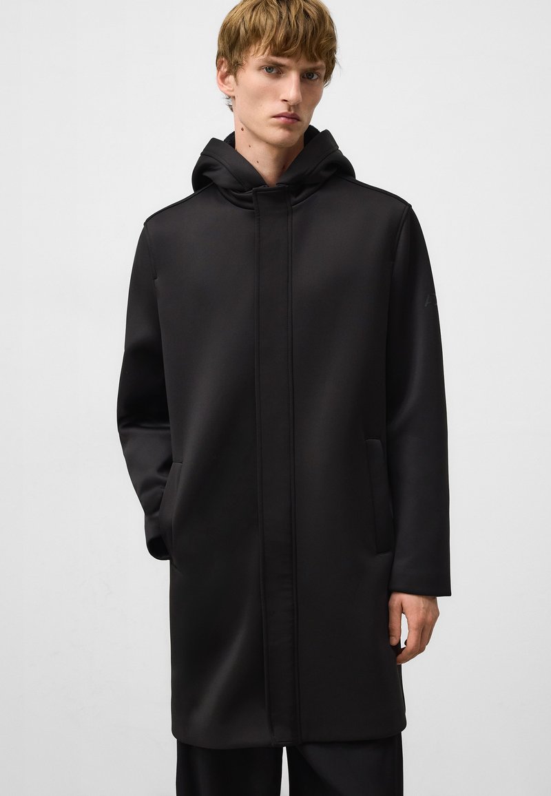 Manteau noir à capuche avec une texture lisse, présentant des poches latérales et un design minimaliste. Le matériau semble imperméable avec des coutures soignées.