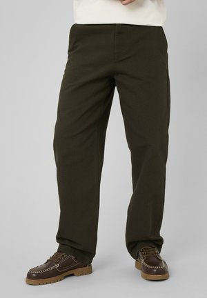 Personne portant un pantalon droit couleur olive foncée et des chaussures marron en cuir à lacets sur un fond gris.