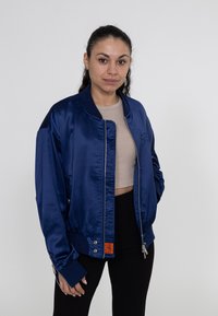 Navy Satin Bomberjacke mit gerippten Bündchen und Saum, ausgestattet mit Seitentaschen und einem orangefarbenen Etikett an der unteren linken Vorderseite.