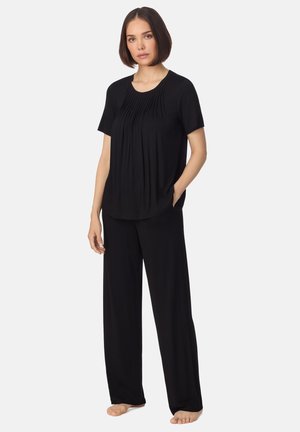 Donna in piedi a piedi nudi, indossa una maglia nera plissettata a maniche corte e pantaloni neri a gamba larga, su uno sfondo chiaro e semplice.