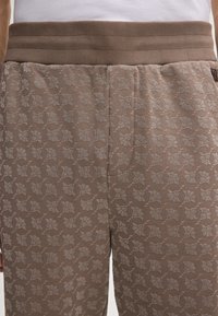 Close-up van lichtbruine broeken met een klein grijs bladerenpatroon en een geribde elastische tailleband, gedragen met een witte top.