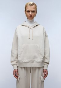 Beige hoodie met een zak aan de voorkant, een geribbelde onderrand en bijpassende beige sweatpants. Het materiaal lijkt een zachte katoenen mix en heeft een relaxte pasvorm.