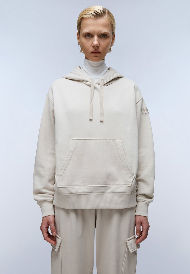 Beige hoodie met een zak aan de voorkant, een geribbelde onderrand en bijpassende beige sweatpants. Het materiaal lijkt een zachte katoenen mix en heeft een relaxte pasvorm.