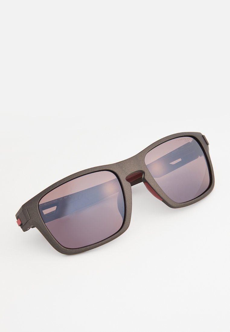 Sonnenbrille mit einem matten braunen Kunststoffrahmen, quadratischen Gläsern in gradientem lila Farbton, roten Akzenten an den Bügeln und einer strukturierten Oberfläche.