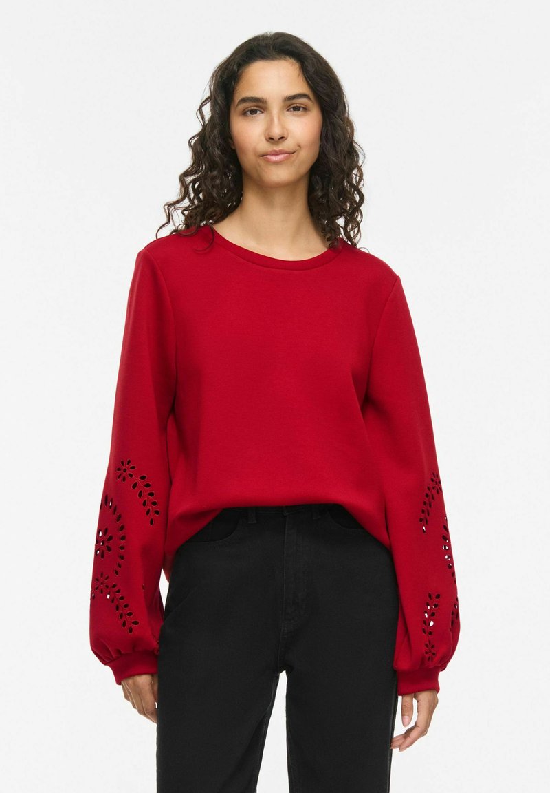 Femme aux cheveux bouclés portant un haut rouge à manches longues avec des motifs floraux découpés sur les manches et un pantalon noir sur fond blanc.