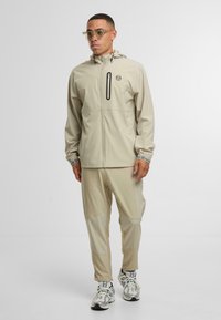 Beżowa kurtka wodoodporna z kapturem, z suwakiem z przodu i logo. W komplecie z pasującymi spodniami z detalami w postaci paneli i subtelną strukturą.