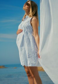 Lichtblauwe en witte gestreepte mouwloze jurk met een vloeiende silhouet, gedragen door een persoon die naast een sheer witte stof staat tegen een zeeachtergrond.