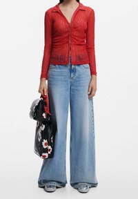 Femme portant un top transparent rouge à manches longues avec boutons, un jean large bleu clair, des chaussures grises, tenant un sac à main noir avec un motif floral blanc et rouge.