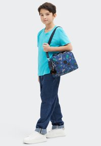 Borsa blu con motivi a tema acquatico, materiale in tessuto, tracolla regolabile e portachiavi in peluche. Indossata con una maglietta turchese e jeans.