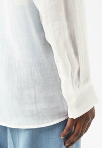 Weißes Leinenhemd mit strukturierter Oberfläche, langen Ärmeln, knöpfbaren Manschetten und lockerer Passform, kombiniert mit hellblauen Denim-Hosen.