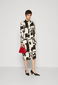 Rochie lungă din material crem cu un model abstract negru, asortată cu pantofi cu vârf ascuțit negri și o geantă mică roșie. Textură netedă a țesăturii.