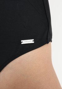Parte inferior de bikini negra hecha de una tela elástica, con una etiqueta de metal plateada que dice "LASCANA" unida en la costura lateral. Textura suave.