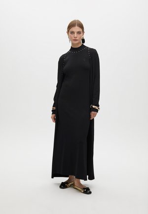 Black long-sleeve dress with a high neckline, embellished with studs. The model wears flat black and gold shoes, showcasing a minimal design.