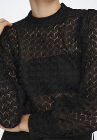 Top in pizzo nero con maniche lunghe, caratterizzato da intricati motivi geometrici, tessuto testurizzato e un design con collo alto. Sezioni trasparenti sono visibili.