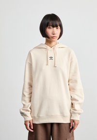 Sweatshirt bege com capuz feito de um tecido macio, apresentando um pequeno logótipo preto "adidas" no peito e mangas de corte relaxado.