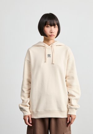 Sweat à capuche beige en tissu doux, avec un petit logo "adidas" noir sur la poitrine et des manches à coupe décontractée.