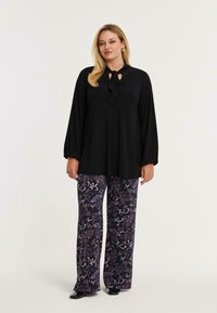 Sorte langærmet bluse med en slips-hals detalje, parret med brede bukser med et lilla blomsterprint på en mørk baggrund.