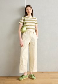 T-shirt rayé olive et crème, pantalon beige à jambes larges, sac en cuir vert, chaussures vertes avec des détails en dentelle, sur un sol en bois clair.