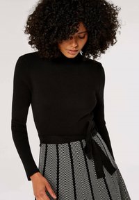 Pull noir à côtes avec des manches longues et un lien à la taille, associé à une jupe à motif chevron noir et blanc.