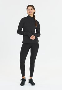 Vrouw staat in een zwarte sportieve zip-up top, zwarte legging en zwarte hardloopschoenen tegen een strak witte achtergrond.