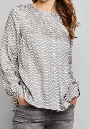 Woman wearing a long-sleeve button-up blouse with a gray and white geometric pattern, hands in pockets, paired with gray jeans.