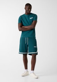 Teal short-sleeve t-shirt with white logo paired with matching shorts featuring white accents and drawstring. Worn with white sneakers.